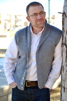 a man standing next to a tree with his hands in his pockets and wearing glasses