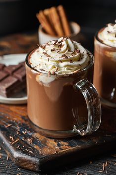 two mugs of hot chocolate with whipped cream on top and cinnamon sticks in the background