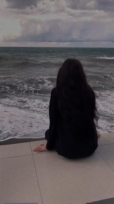 a woman sitting on the edge of a pier looking out at the ocean and storm clouds