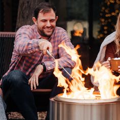 two people sitting around a fire pit while one holds a drink and the other eats