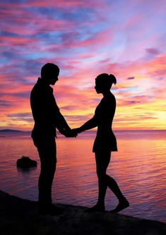 two people holding hands while standing on the beach in front of an orange and purple sunset