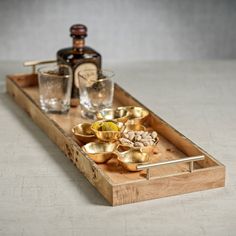 a wooden tray with gold bowls and glasses on it next to a bottle of alcohol