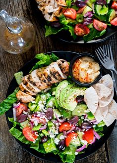 two black plates filled with salad and chicken on top of a wooden table next to a glass of water