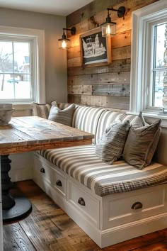 a wooden bench sitting under two windows next to a table with pillows on top of it