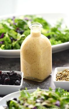 salad dressing in a glass bottle surrounded by bowls of greens, raisins and sunflower seeds