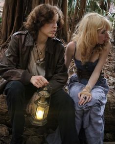 two people sitting next to each other in the woods with a lantern on their lap