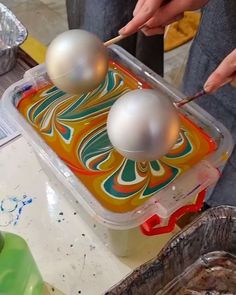 two balls are in a plastic container on a table with other items around it and one is holding a paintbrush