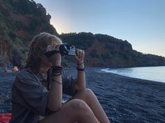 a woman sitting on top of a beach holding a camera up to her face while looking into the distance