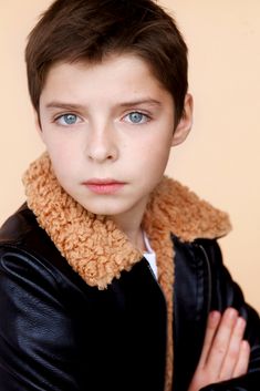 a young boy wearing a black leather jacket with a brown collar around his neck and hands on his chest