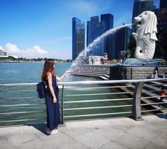 a woman standing next to a water fountain near the ocean with buildings in the background