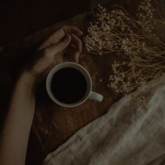 a person holding a cup of coffee on top of a wooden table next to dried flowers