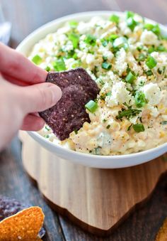 a person dipping a tortilla chip into a bowl of dip with cheese and green onions