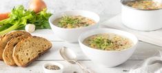 three bowls of soup are next to some bread and vegetables on a white wooden table