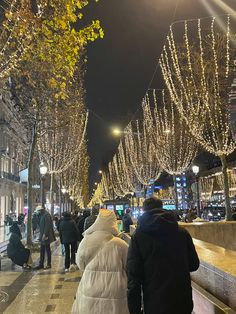 people walking down the street at night with christmas lights on trees and buildings in the background