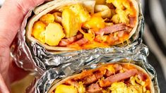 two breakfast burritos wrapped in foil with scrambled eggs and bacon on them, being held by someone's hand