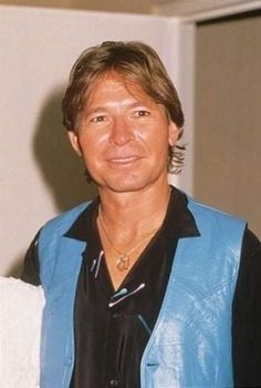 a man wearing a blue vest and black shirt standing in front of a white wall