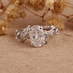 a close up of a ring on a table with flowers in the background and an oval shaped diamond surrounded by vines