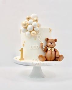 a teddy bear sitting on top of a white cake with gold balloons attached to it