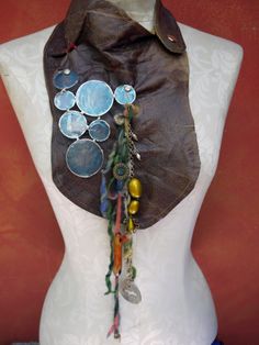 This cool bib collection is mostly recycled! The leather is remnants from a furniture maker, the blue capiz circles are repurposed from a candle holder and the brass hardware is from vintage jewelry. I cinched the leather creases with thick red thread and connected the shells with screw head anchors. The colorful yarn was hand spun suspending cast concrete charm.Â  Finished with gold top stitching Measures 10" long,(16" with fringe) 8" wide Neck measures 13" around Cast Concrete, Recycle Bin, Red Thread, Leather Fringe, Bib Necklace