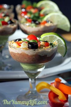 two glasses filled with food and garnished with limes, tomatoes, peppers, olives, and other vegetables