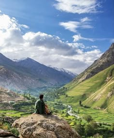 a man sitting on top of a large rock in the middle of a green valley