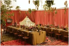 a long table is set up with flowers and greenery in front of an orange drapes