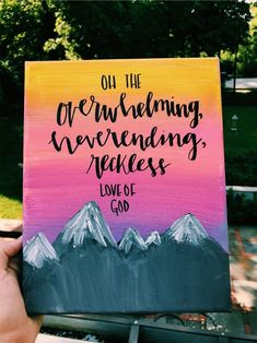 a hand holding up a painting with mountains in the background and bible verse on it