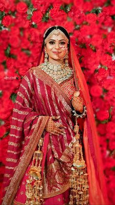 a woman in a red and gold bridal outfit posing for the camera with flowers behind her