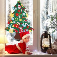 a baby wearing a santa claus outfit sitting in front of a window next to a christmas tree