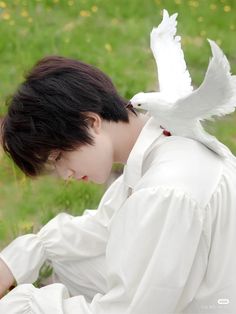 a young man sitting in the grass with a white bird on his shoulder