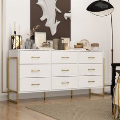 a white dresser with gold handles and drawers in a living room next to a painting on the wall