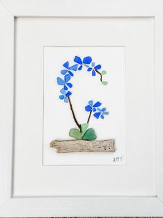 a white frame with blue flowers and green leaves on it, sitting next to a piece of driftwood