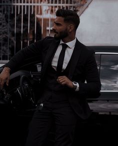 a man in a suit and tie standing next to a car with his hand on the steering wheel