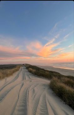 a dirt road that is next to some grass and sand with the sun setting in the background