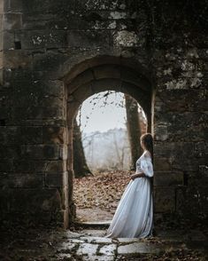 a woman in a long white dress is standing by an old brick wall and looking into the distance