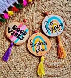 three hand painted wooden tags with tassels on top of a woven mat, one saying good vibe be happy and the other saying kindness is free