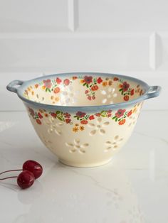 a white bowl with red and yellow flowers on it next to a cherries sprig