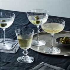 three wine glasses with olives in them sitting on a silver platter next to a bowl of green olives