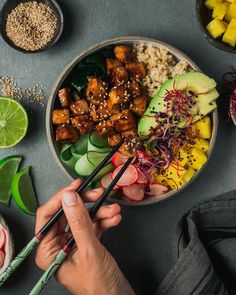 someone holding chopsticks over a bowl of food with rice, avocado and tofu