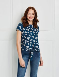 a woman standing in front of a white wall wearing jeans and a top with flowers on it