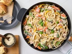 a skillet filled with pasta and vegetables on top of a table next to bread