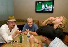 three men sitting at a table with drinks and food in front of them, laughing