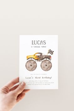 a hand holding up a card with a monster truck on it and the words lucas is turning three