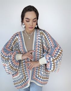 a woman wearing a multicolored crocheted cardigan sweater and jeans standing in front of a white wall