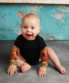 a baby sitting on the floor with his arms and legs covered in tattoo sleeves, smiling at the camera