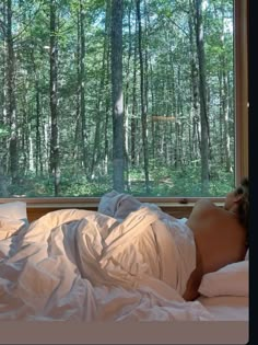 a naked woman laying in bed next to a window looking out at the woods outside