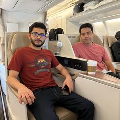 two men sitting on an airplane next to each other with drinks in their hands and looking at the camera