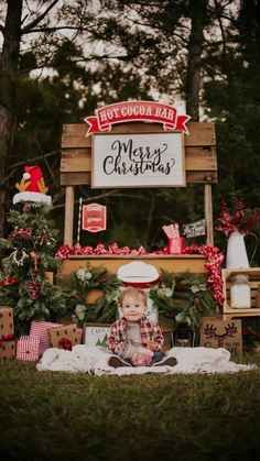 a baby sitting on the ground in front of a merry christmas sign with presents around it