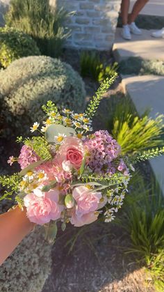 a hand holding a bouquet of pink flowers and greenery in front of a brick wall