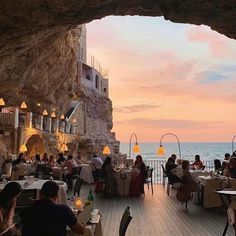 people are sitting at tables in an outdoor restaurant overlooking the ocean and buildings with lights on them